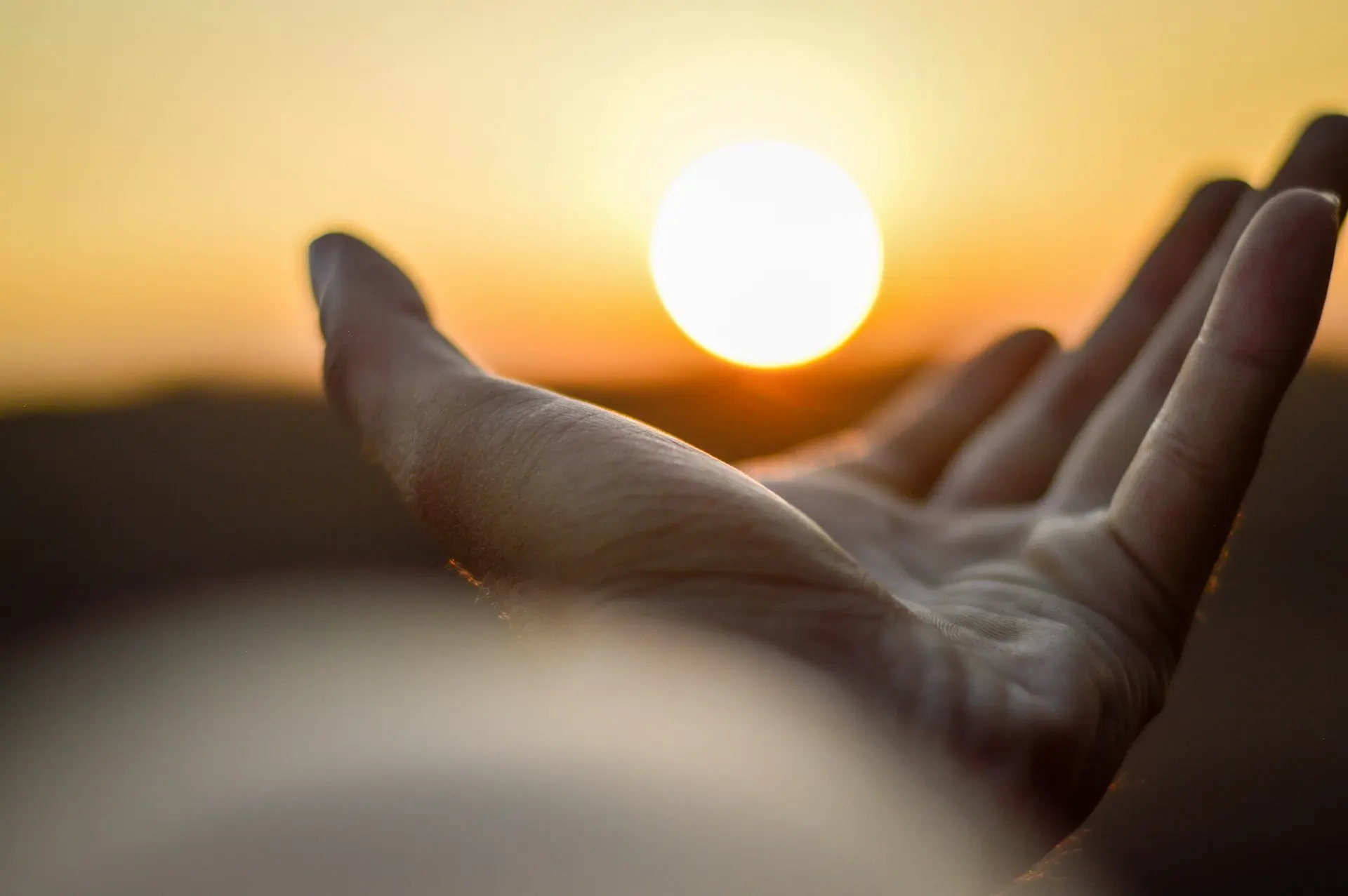 An open human hand reaching toward a bright low sun in a warm sky
